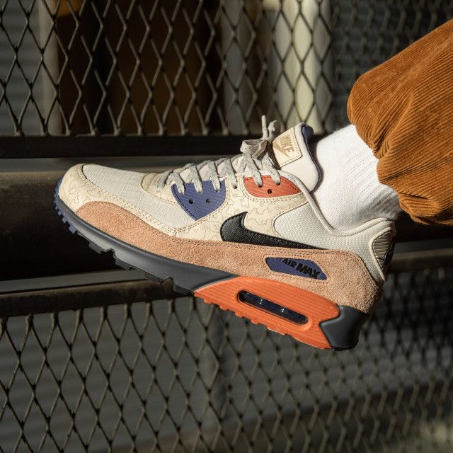 In hand shot van de Camowabb Air Max 90 met zicht op suède toebox, oranje tonglabel en camoprint – premium texturen en ACG-style design