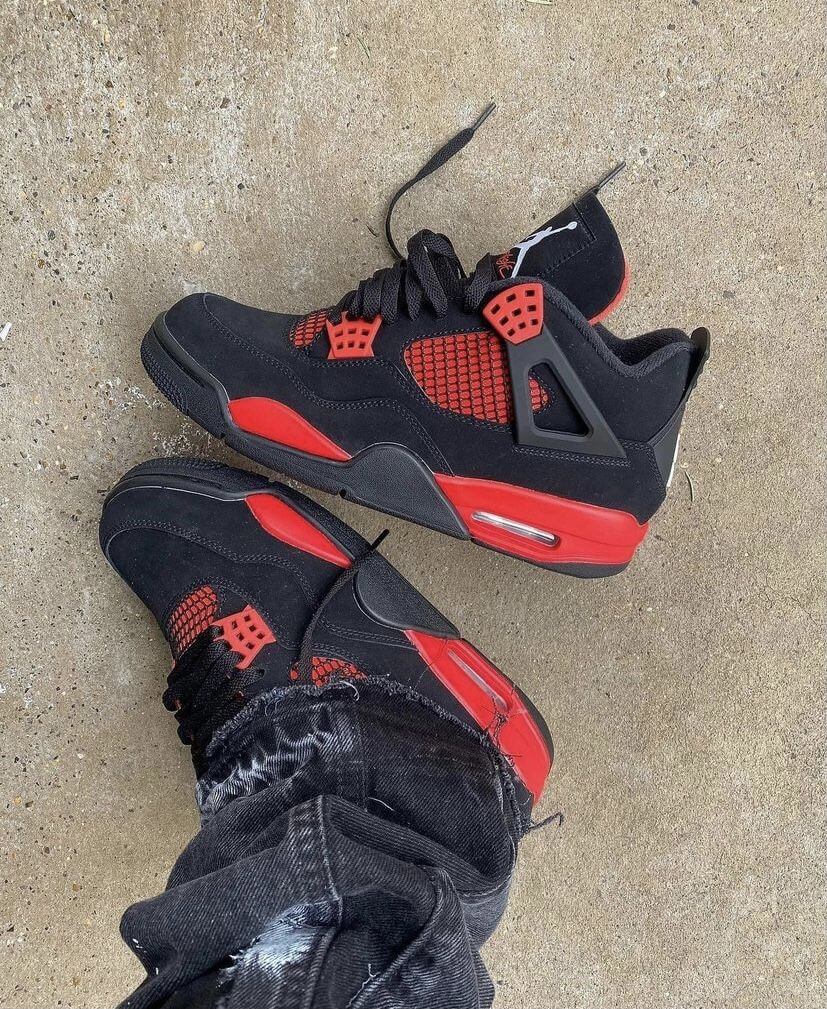Jordan 4 Red Thunder CT8527-016 half uit doos geschoven met zicht op het matte zwart suède en de dieprode tonginzet onder kunstlicht
