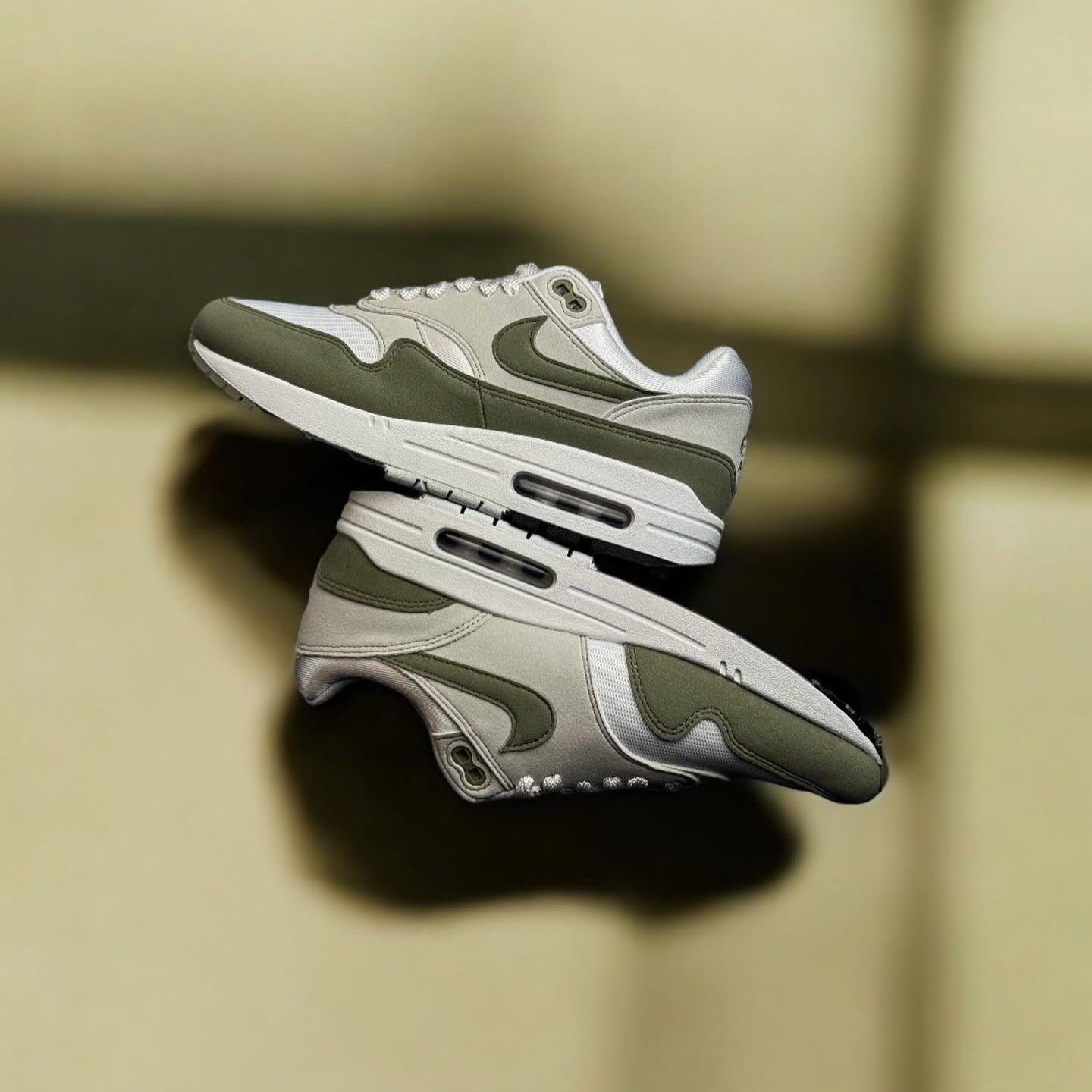 Drie paren Air Max 1 Light Army zwevend gestyled tegen zachte achtergrond, focus op tonale balans en retro-runner shapes