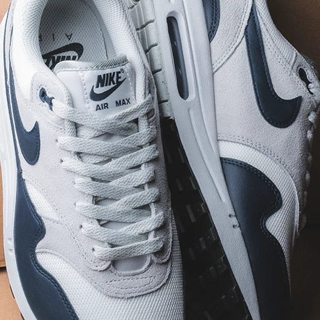 Sneakers strak gepositioneerd tegen beige achtergrond met nadruk op de gewatteerde tong, zichtbare Air-unit en donkere toebox