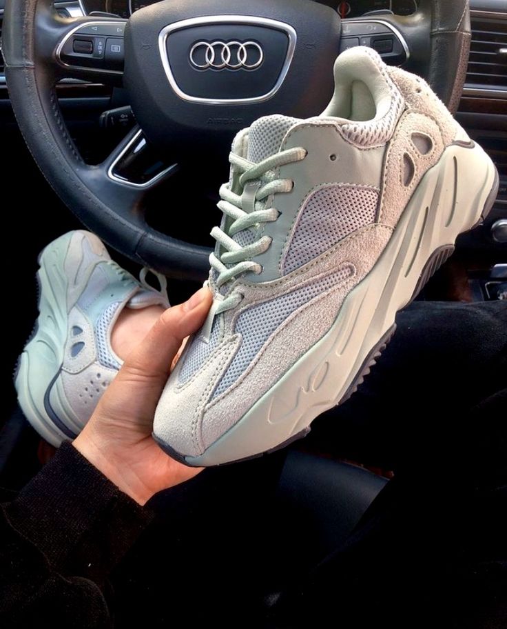 Close-up van de Adidas Yeezy 700 in lichtgrijze tinten vastgehouden in een auto interieur met Audi stuur, waarbij de chunky zool en layered panelen duidelijk zichtbaar zijn