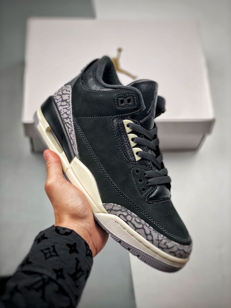 Hand houdt een paar Air Jordan 3 Off Noir omhoog in retailsetting met focus op details en textuur