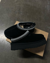 UGG Tazz Slipper Black 1122553-BLK geplaatst op UGG-doos, zwarte suède upper en contrasterende gevlochten kraag duidelijk zichtbaar in zachte studioverlichting, vastgelegd door Snrkickz.