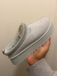 Close-up van UGG Tazz Slipper Goose 1122553-GOO, lichtgrijs suède met gevlochten rand en stevige platformzool, vastgehouden voor detailweergave door Snrkickz.