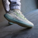 Lifestyle opname van de Yeezy Boost 380 Alien Blue gedragen in urban setting, waarbij de blauwe zool en groene details contrasteren met een betonnen vloer voor trendy streetwear vibe