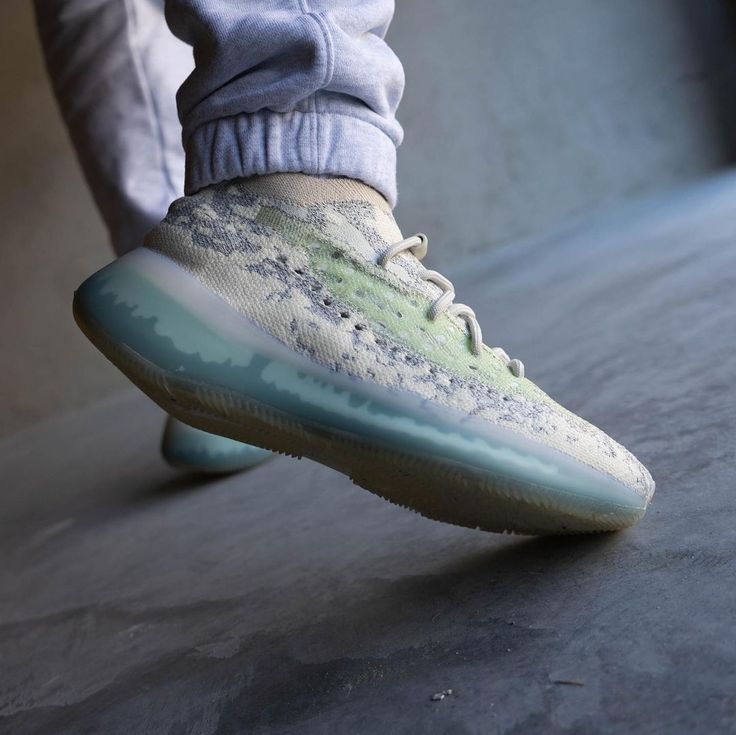 Lifestyle opname van de Yeezy Boost 380 Alien Blue gedragen in urban setting, waarbij de blauwe zool en groene details contrasteren met een betonnen vloer voor trendy streetwear vibe