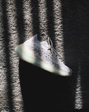 rtistieke opname van de Yeezy Boost 380 Calcite Glow met licht en schaduwpatronen, waarbij de glow-in-the-dark zool en futuristische details goed naar voren komen