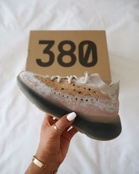 Lifestylebeeld van Yeezy 380 Pepper boven wit bed, vastgehouden met de box voor mobile shoppers die zoeken op 'yeezy pepper real-life look' of 'sneaker styling'