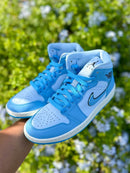 In-hand shot van Air Jordan 1 Mid Ice Blue – zachte blauwe panelen, witte zool en zwarte branding in daglicht zichtbaar