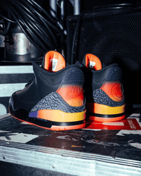  Close-up van de hielen van de J Balvin Jordan 3 met regenboogkleurige smiley-logo's en feloranje outsole, gefotografeerd in industriële setting