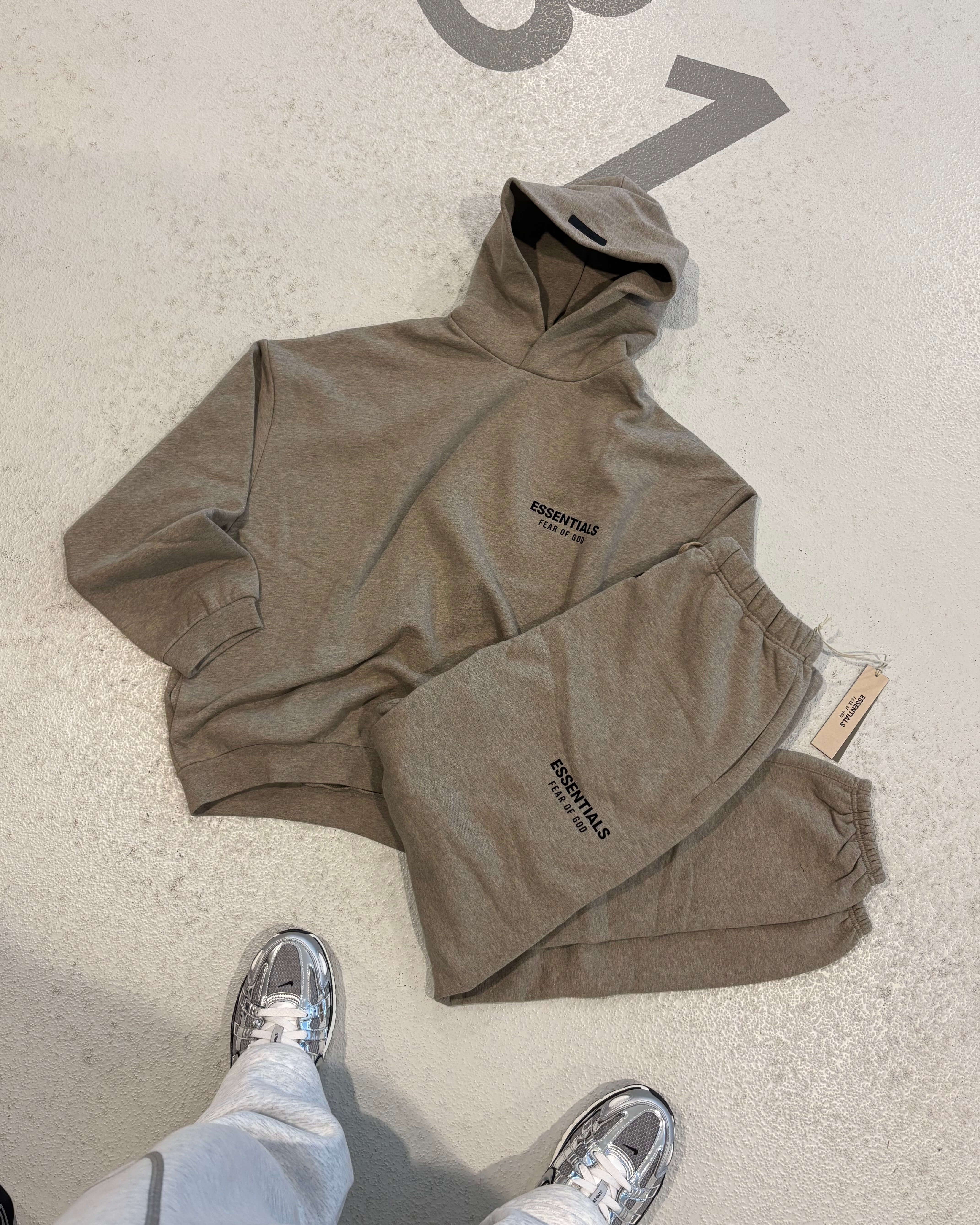 FFear of God Essentials FW24 Light Heather sweatpants flat lay showing elastic cuffs, Essentials branding and beige drawcords. In-hand view of Fear of God Essentials FW24 Light Heather set showing soft cotton texture and relaxed streetwear style.