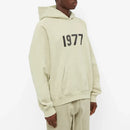 Model wearing Fear of God Essentials 1977 Wheat Hoodie highlighting oversized fit, neutral beige shade and signature 1977 chest print.
