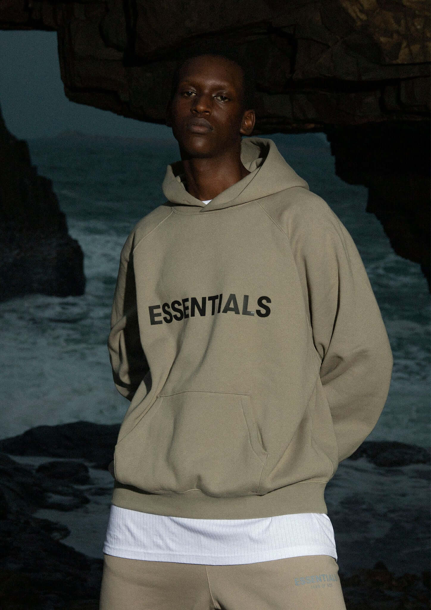 Fear of God Essentials Hoodie Moss – model draagt mosgroene hoodie bij rotsachtige kustlijn in outdoor setting