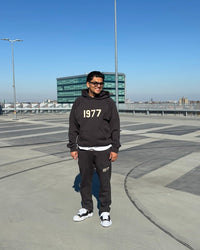 Close-up van de Fear of God Essentials IRON 1977 hoodie en pants gedragen in een open ruimte, met zichtbare oversized fit