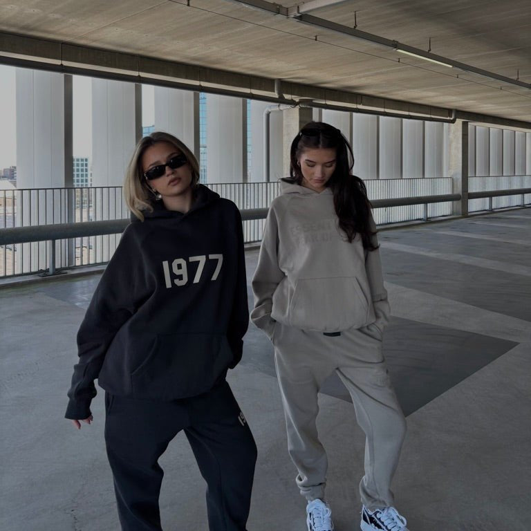 Twee personen dragen de Fear of God Essentials IRON 1977 Full Set buiten in urban omgeving, gestyled met sneakers