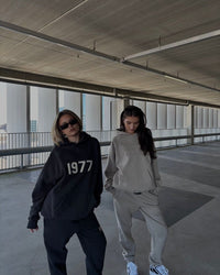 Twee personen dragen de Fear of God Essentials IRON 1977 Full Set buiten in urban omgeving, gestyled met sneakers