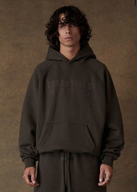 Model draagt de Fear of God Essentials Off Black SS22 Full Set, bestaande uit hoodie en joggers in diepe Off Black kleur, relaxed fit van zacht katoen met subtiel Essentials-logo voor een moderne streetwear look