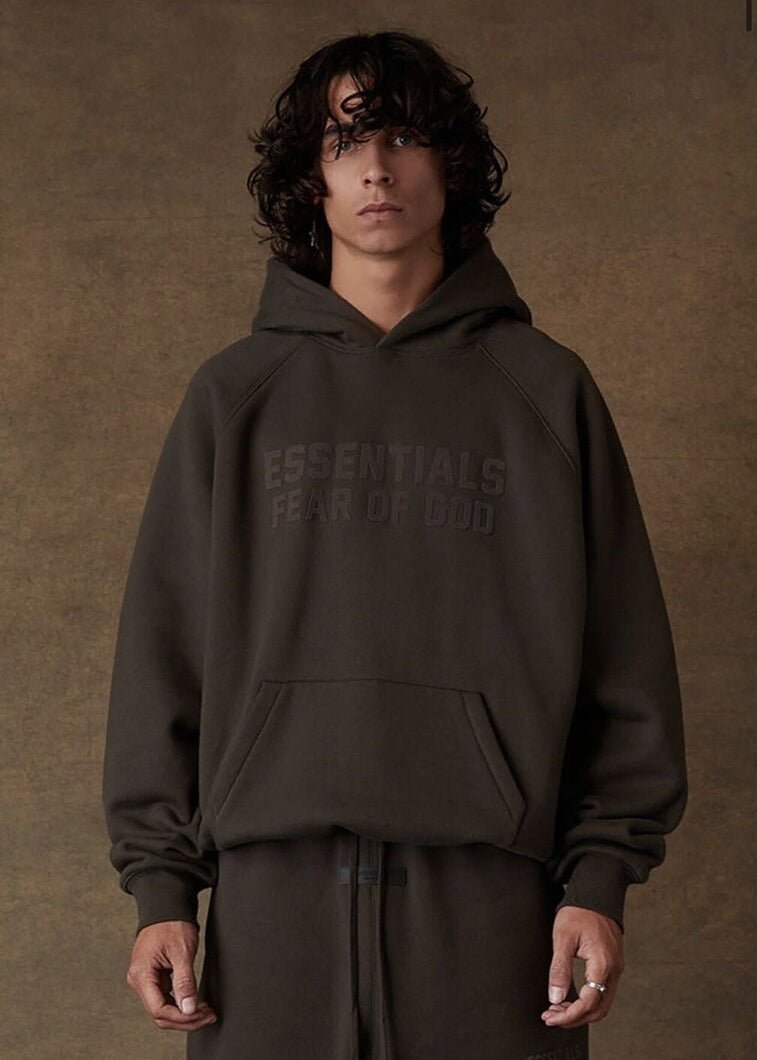 Model draagt de Fear of God Essentials Off Black SS22 Full Set, bestaande uit hoodie en joggers in diepe Off Black kleur, relaxed fit van zacht katoen met subtiel Essentials-logo voor een moderne streetwear look