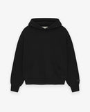 Model wearing Fear of God Essentials SS25 Jet Black Hoodie highlighting oversized fit, tonal logo print and clean modern streetwear style.