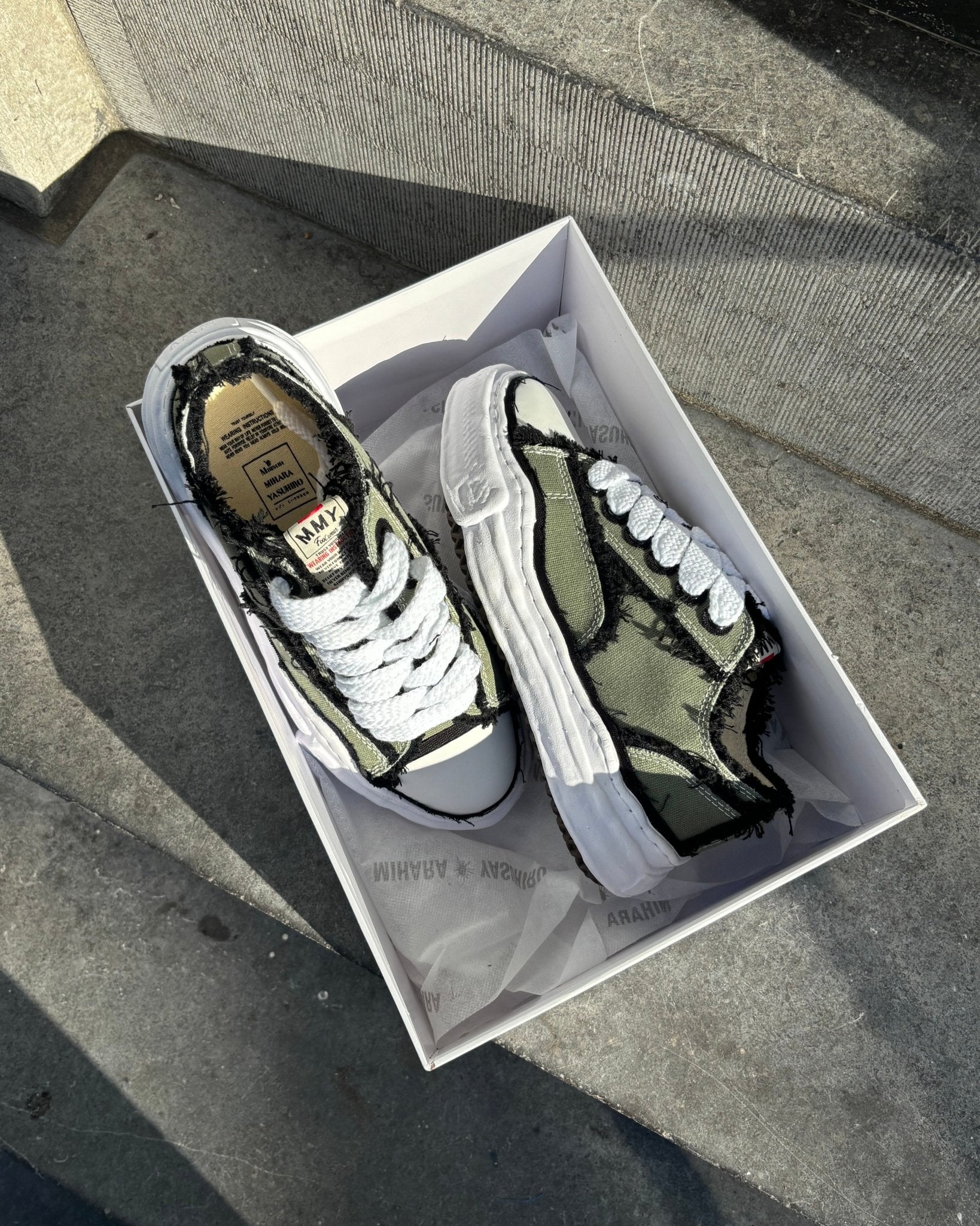 Maison Mihara OG Sole Low Khaki in open doos op stoep, natuurlijke schaduwen benadrukken het ruwe canvas en dikke zool