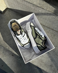 Maison Mihara OG Sole Low Khaki in open doos op stoep, natuurlijke schaduwen benadrukken het ruwe canvas en dikke zool