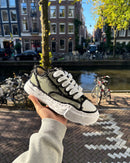 Maison Mihara OG Sole Khaki sneaker vastgehouden in Amsterdam met grachtenpand en fietsen op de achtergrond, typisch Snrkickz straatbeeld