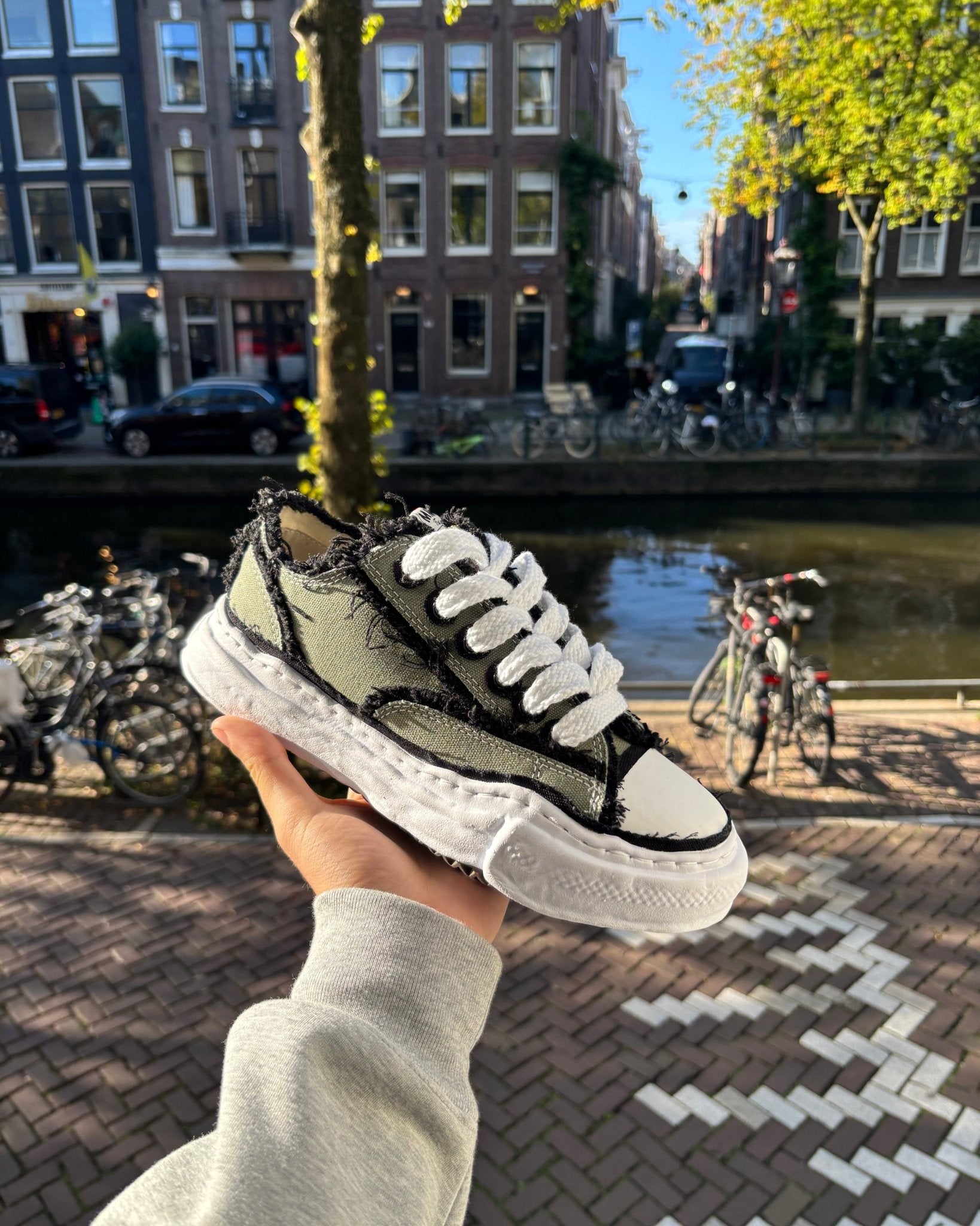 Maison Mihara OG Sole Khaki sneaker vastgehouden in Amsterdam met grachtenpand en fietsen op de achtergrond, typisch Snrkickz straatbeeld