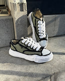 Maison Mihara OG Sole Low Khaki zijaanzicht geplaatst op traptrede, met sterke focus op golvende zool en ruwe canvastextuur