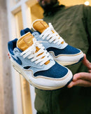 Nike Air Max 1 Denim Aura opgetild bij een raam, met gele tonglabel en bovenwerk dat overloopt van washed blue naar zandkleurige tinten