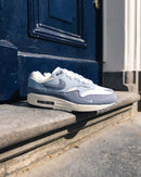 Nike Air Max 1 Light Armory Blue op stoeprand geplaatst bij Amsterdamse voordeur, classic Air Max styling in urban setting