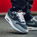 In-hand shot van de Air Max 1 ’87 Dark Smoke Grey met ruwe textuur, zwarte accenten en zichtbare Air-unit
