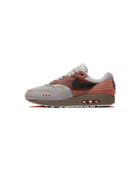 Zijaanzicht van de Air Max 1 Amsterdam met baksteenrode tinten, suède overlays en een minimalistische, cleane achtergrond die het silhouet versterkt