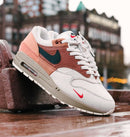 Urban shot met reflectie op natte stoeptegels, waarin de Air Max 1 Amsterdam de rode accenten en het stadsgevoel van de colorway versterkt