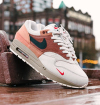 Urban shot met reflectie op natte stoeptegels, waarin de Air Max 1 Amsterdam de rode accenten en het stadsgevoel van de colorway versterkt