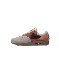 Zijaanzicht van de Air Max 1 Amsterdam met baksteenrode tinten, suède overlays en een minimalistische, cleane achtergrond die het silhouet versterkt