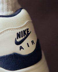 Hielshot van de Nike Air Max 1 in Coconut Milk met navy hielpaneel, geborduurde branding en stevige suède constructie