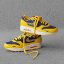 Sfeerbeeld van de sneaker in gedempt licht op betonnen vloer, waarbij de gele tonglabel en navy Swoosh sterk contrasteren