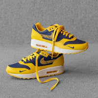 Sfeerbeeld van de sneaker in gedempt licht op betonnen vloer, waarbij de gele tonglabel en navy Swoosh sterk contrasteren