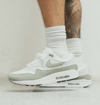 Sneaker zichtbaar aan voet met sokken, tegen beige vloer, met lichte beweging en schaduwspel dat het silhouet versterkt