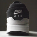 In hand shot van de Nike AM1 Off Noir Vast Grey met zacht suède, offwhite midsole en stevige constructie