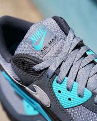 In hand shot van de Nike AM90 Cool Grey & Dusty Cactus met zachte materialenmix, witte zool en contrasterende turquoise highlights