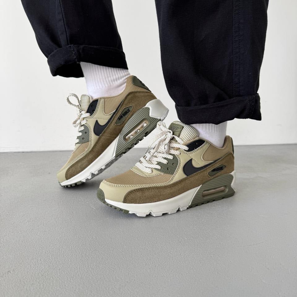 In hand shot van de Nike AM90 Neutral Olive met canvas textuur, suède mudguard en licht crèmekleurige zool
