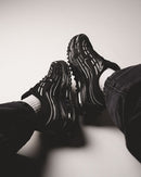 Lifestylebeeld van Nike Air Max TL 2.5 Black Metallic Silver op betonnen vloer, beide schoenen diagonaal geplaatst met focus op de zoolconstructie