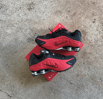 Twee Nike Shox R4 sneakers gestyled naast elkaar op ruwe betonnen ondergrond, met focus op rode kleur en reflectie