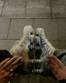 Handen houden de Nike Shox TL Chrome vast onder grauwe TL-verlichting, met focus op de metallic upper en reflectie