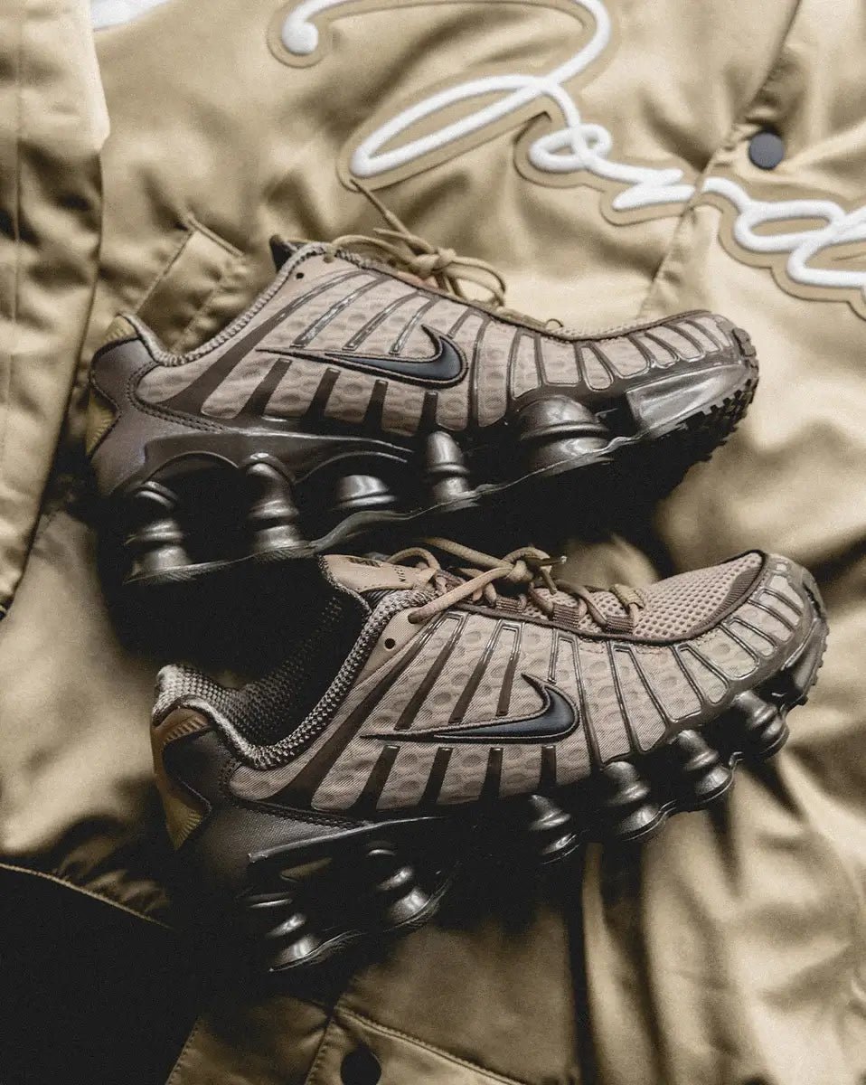 Nike Shox TL Khaki Ironstone Off Noir als paar gefotografeerd liggend op een khaki achtergrond, met nadruk op de earthy kleuren en layered details