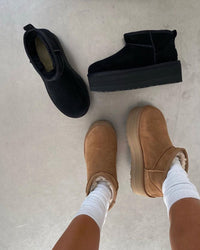 In-hand beeld met contrast tussen zwarte suède UGGs en beige vloer, voeten van model in witte sokken en camel slippers op achtergrond