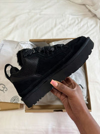 De zwarte UGG Lowmel geplaatst op een wit dekbed, vastgehouden met één hand, benadrukt cozy styling