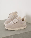 UGG Lowmel Ceramic 1144032-CERA in zachte beige tint, geplaatst op betonoppervlak in lifestyle setting met zichtbare fleece details en suède textuur, vastgelegd door Snrkickz.