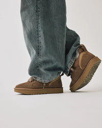 On-feet foto van UGG Lowmel Hickory gecombineerd met wijdvallende jeans, gestyled in studio met witte achtergrond